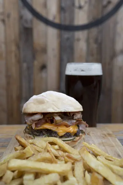 Monster burger. View of a giant burger with bread, meat, cheddar cheese, onions, bacon, cucumber pickles, sauce, french fries and a pint of black beer on the restaurant wooden table.