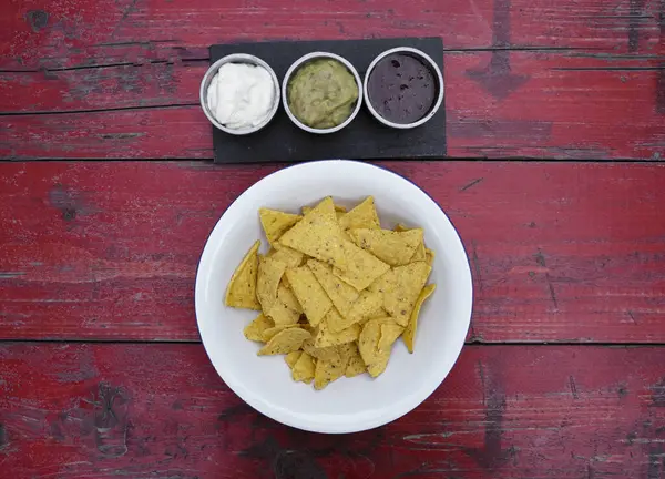 Meksika yemeği. Beyaz kasedeki nachos 'un üst görüntüsü ve kırmızı ahşap masada üç soslu çedar peyniri, acı biber ve guacamole..