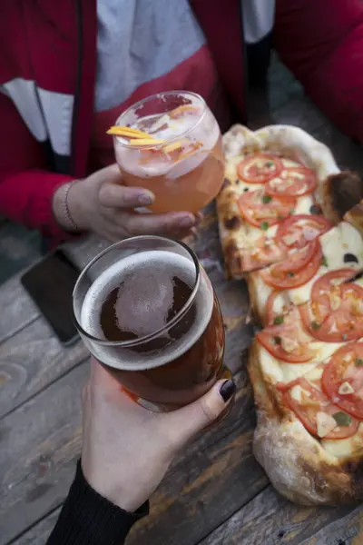 Having pizza and beer at the restaurant. Closeup view of a man and woman toasting with beer and a tropical cocktail while dining a traditional pizza with tomato, garlic and oregano.