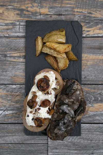 Gourmet sandwich. Top view of a grilled veal steak sandwich with fresh and grilles tomatoes, almonds and burrata cheese, with roasted potatoes as garnish, served in a black dish on the wooden table.