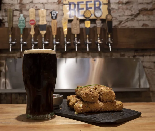 View of a glass of black beer and fried mozzarella sticks in a black dish on the wooden restaurant counter.