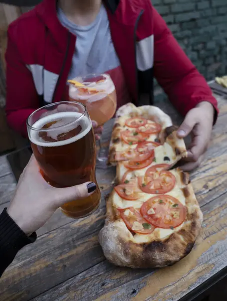 Having pizza and beer at the restaurant. Closeup view of a man and woman toasting with beer and a tropical cocktail while dining a traditional pizza with tomato, garlic and oregano.