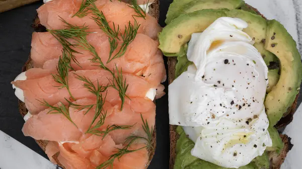 Healthy cuisine. Gourmet sandwich. Closeup view of a smoked salmon, cream cheese, sliced avocado and poached eggs sandwich with fried potatoes on a black dish on the white marble table.