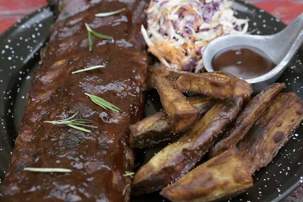 Barbekü sosu ve otlarla marine edilmiş domuz pirzolası, patates kızartması ve lahana salatası, kırmızı ahşap masada siyah bir tabakta.. 