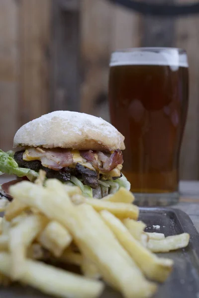 View of a traditional burger with bread, bacon, cheddar cheese, lettuce and tomato, french fries and an amber ale beer on the wooden table.