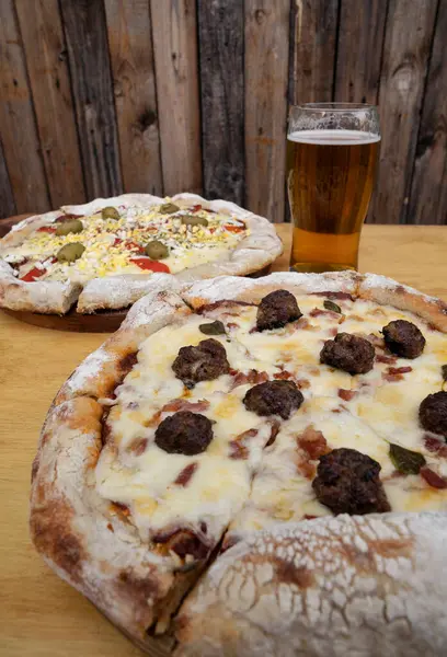 Eating at the restaurant. Closeup view of a pizza with mozzarella cheese, oregano, bacon and meatballs, with a pint of beer. Another pizza with cheese, egg, peppers and ham in the background.
