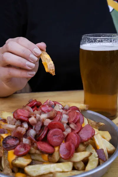 Closeup view of a woman holding a fried potato dipped in cheddar cheese sauce. The french fries with sausages and bacon dish besides a glass of beer.
