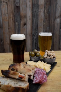 Arjantin Picada 'sı. Peynirli, salamlı, fıstıklı, yeşil zeytinli ve Focaccia ekmekli, iki bardak biralı ve ahşap arka planlı.