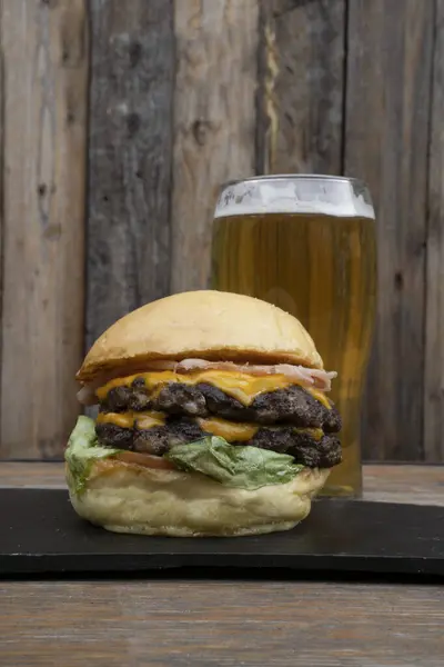 Food and drink. Closeup view of a multilayer burger with bread, lettuce, tomato, cheddar cheese and ham, and a pint of beer, on the restaurant wooden table with a wooden background.