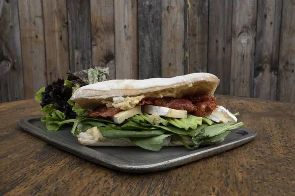 Gurme sandviç. Ciabatta ekmeği, nohut humusu, avokado, kurutulmuş domates, brie peyniri ve ıspanaklı vejetaryen sandviçin ahşap masada metal bir tabakta yakın görüntüsü.. 