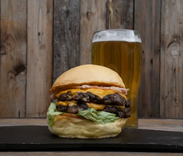 Food and drink. Closeup view of a multilayer burger with bread, lettuce, tomato, cheddar cheese and ham, and a pint of beer, on the restaurant wooden table with a wooden background.