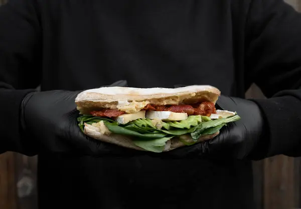 Gourmet sandwich. Closeup view of the chef wearing black gloves, holding a vegetarian sandwich with ciabatta bread, chickpea hummus, avocado, sun dried tomatoes, brie cheese and spinach.