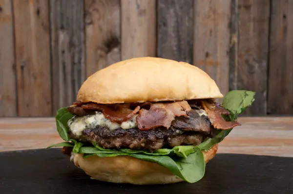 Delicious burger with bread, bacon, mozzarella, blue cheese, meat and spinach leaves, with a wooden background.