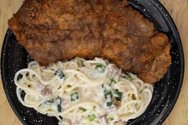 Pasta and meat. Top view of a milanesa, breaded steak, and spaghetti with a bacon and leek cream sauce, in a black dish on the restaurant wooden table.