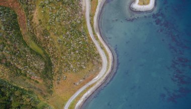 Doğal hava fotoğrafçılığı. Okyanus kıyısından başka toprak yolun en üst görüntüsü, yeşil tarla ve ormanı turkuaz deniz suyundan ayırıyor.