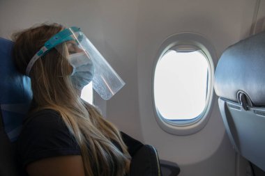 Koronavirüs salgını sırasında seyahat etmek. Uçağın içinde, koruyucu bir maske takmış genç bir kadının görüntüsü.. 