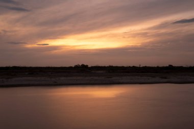 Akşamüstü nehrin uzun pozlu görüntüsü. Su yüzeyindeki gökyüzünün yansımasının görüntüsü. Manzarada güzel sarı ve turuncu günbatımı renkleri.