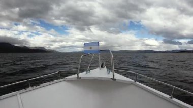 Bulutlu görkemli bir gökyüzünün altında Beagle Kanalı 'nı geçmek. Geminin pruvasında dalgalanan Arjantin bayrağı ve kıyıdaki And Dağları manzarası