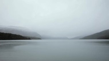 Göle düşen yağmur damlaları. Yağmurlu bir sabahta bulutlu beyaz gökyüzü ile buzullu göl, orman ve dağların manzarası.