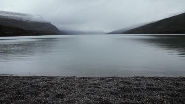 Gölde yağmur yağıyor. Sakin göl manzarası, bulutlu bir gökyüzünün altındaki tepeler ve orman. Su damlaları gölün yüzeyine düşer.