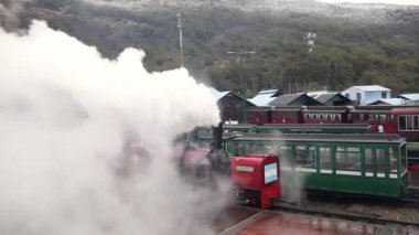 Tierra del Fuego, Arjantin 04,25,2025: Tren yolu Austral Fueguino kırmızı lokomotif ve vagonlar istasyondan ayrılıyor. Lokomotiften beyaz duman çıkıyor. Orman ve tepeler arka planda
