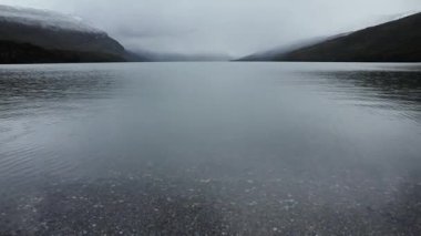 Gölde yağmur yağıyor. Sakin göl manzarası, bulutlu bir gökyüzünün altındaki tepeler ve orman. Su damlaları gölün yüzeyine düşer.. 