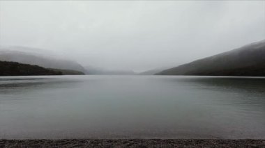 Gölde yağmur yağıyor. Sakin göl manzarası, bulutlu bir gökyüzünün altındaki tepeler ve orman. Su damlaları gölün yüzeyine düşer.
