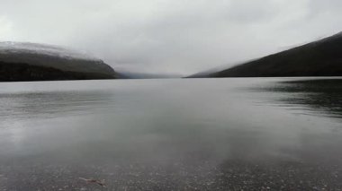 Gölde yağmur yağıyor. Sakin göl manzarası, bulutlu bir gökyüzünün altındaki tepeler ve orman. Su damlaları gölün yüzeyine düşer.