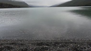 Gölde yağmur yağıyor. Sakin göl manzarası, bulutlu bir gökyüzünün altındaki tepeler ve orman. Su damlaları gölün yüzeyine düşer.