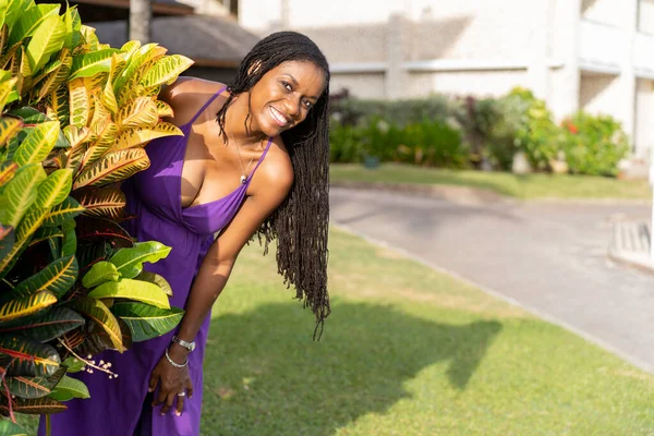Black woman with long natural hair smiling happily peeking out from behind a croton plant