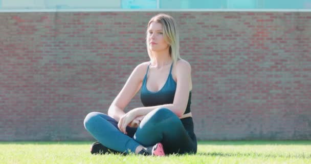 スポーツ黒と青の上に長いブロンドの髪を持つ若いスポーティーな女の子をフィットさせ レギンスは草や笑顔に座っています 高品質4K映像 — ストック動画