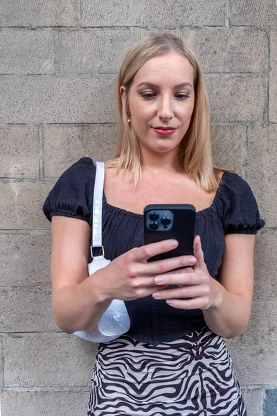 Blonde Young Woman Looks Her Smartphone Smiles She Wears White — Stock Photo, Image