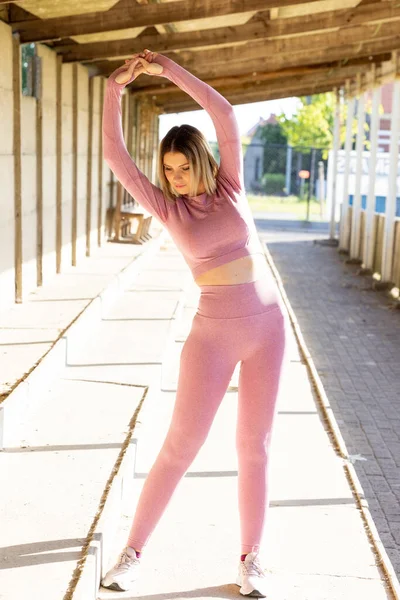 Full body image of a Beautiful young blonde fitness woman wearing pink sports clothes stretching and full body warming up in morning. Sporty woman doing warm up workout by a wall. High quality photo