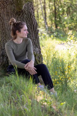 Güneşli bir günde, ormanın içindeki bir ağacın altında çimlerde oturup rahatlayan şık bir esmer. Sağlıklı yaşam tarzı ve rahatlama. Yüksek kalite fotoğraf