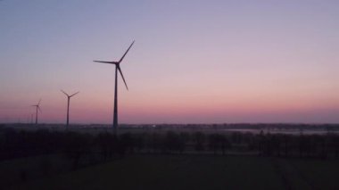 Drone, güneş doğarken kırsal bir arazide sis eşliğinde rüzgar türbini çiftliği vurdu. Yakın çekim rüzgar türbinleri gün doğumunda. Yel değirmenleri yeşil enerji üretiyor. Büyük rüzgâr türbinleri, parlak turuncu pervaneli.