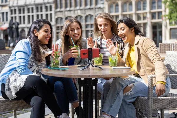 stock image Four multi ethnic female friends, tourists or students sitting at a cafe terrace in the city center using mobile phone for a video call. High quality photo