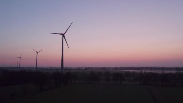日の出や日没の背景に対して緑のフィールドの間で回転翼を持つ風車の無人機によって撮影されました 風力発電用のタービンケーブル付きの風力発電所 再生可能エネルギー源 地球ケア — ストック動画