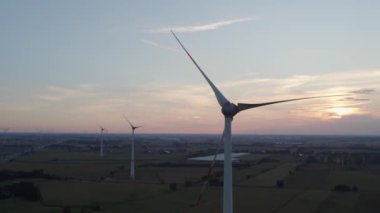 Yeşil alanlar arasında dönen kanatlı yel değirmenleri tarafından gündoğumu ya da günbatımı arka planına karşı çekilen hava görüntüsü. Rüzgar enerjisi için türbin kabloları olan rüzgar çiftliği. Yenilenebilir enerji kaynağı, toprak bakımı