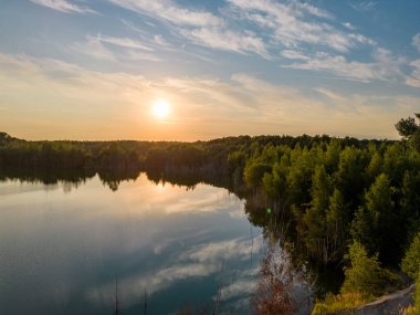 Barışçıl orman gölünün kıyısında, dramatik ve renkli günbatımında turuncu, sarı ve gri bulutlu ağaçların insansız hava aracı görüntüleri. Yüksek kalite fotoğraf
