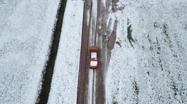 Hava görüntüsü, turuncu bir arabanın arkasındaki dron tarlalar arasında karla kaplı toprak bir yoldan geçiyor. Yüksek kalite 4k görüntü