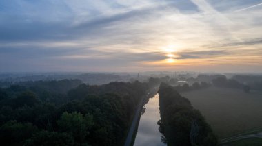 Güneşin doğuşunu ya da batışını gösteren bir dron tarafından çekilen hava görüntüsü ve ağaçları sıcak akşam ışığı ile dolduran sis, altın ışık ışınlarıyla yükseliyor. Bulutlar ve bulutlar yansıyor