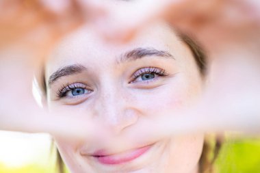Doğada gülümseyen bir kadının ellerle kalp şeklini oluşturduğu görüntüsünü kapat. Dışarıdaki kameraya gülen oldukça neşeli beyaz kadın. Sağlıklı yaşam tarzı, kendini sevme ve vücut bakımı konsepti. Yüksek