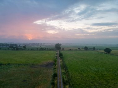 Sise giden dolambaçlı bir kır yolunun merkez odak noktasını oluşturduğu yaz manzarasının nefes kesici bir görüntüsü. Dramatik bir gün batımına karşı çekilen fotoğraf ruhani bir ahenk yaratır.