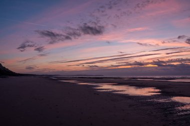 Kumlu sahilde ve denizin ritmik dalgalarında yansıtılan dramatik bir günbatımı görüntüsü. Renklerin nefes kesen oyunu gerçekten büyüleyici bir manzara yaratıyor.