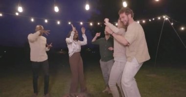 Uyumlu Kutlama: Akşam Bahçesinin Işıkları Altında Dans Eden Çok Kültürlü Arkadaşlar, bir bahçe partisinde ışıkların altında dans eden bir grup etnik arkadaş. Yüksek kalite 4k