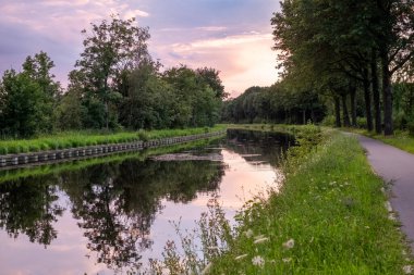 Mor dramatik bir günbatımının nefes kesici bir sahnesi gökyüzünü zengin renklerle boyuyor, sakin sularda güzel bir şekilde yansıtılıyor bereketli ağaçlar arasında zarif bir şekilde akan bir kanal. Büyüleyici.