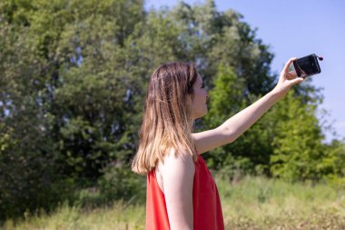 Canlı kırmızı bir etekle süslenmiş güzel beyaz bir kadının cep telefonuyla yemyeşil orman gölleri arasında mükemmel bir resim yakaladığı resim gibi bir sahne. Güneşler
