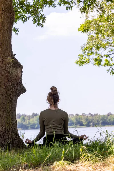 A serene and tranquil moment captured in nature, featuring a young brunette woman sitting gracefully in the lush green grass next to a majestic tree. She faces the peaceful forest lake, her eyes