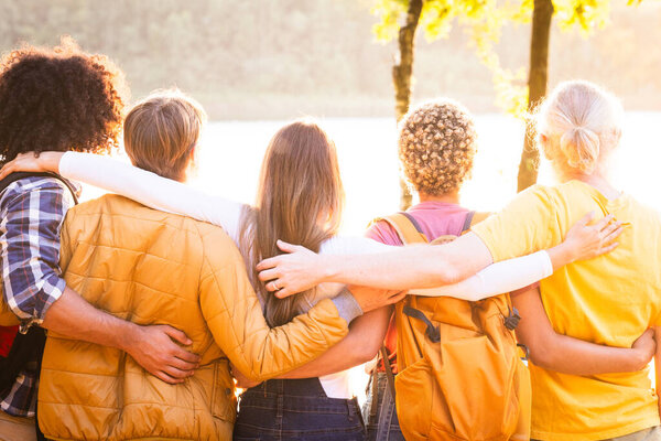 Дружба, многонациональность, единение, путешествия, отдых, праздники, единение, осмотр достопримечательностей. Молодые люди с рюкзаками стоя крепко обнимаясь любуясь естественным видом заката над