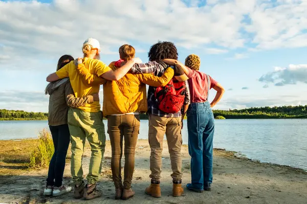 Wanderlust lifestyle, Back view of group of young multi racial friends camping standing hugging on shoulder together on the shore of a forest lake. High quality photo